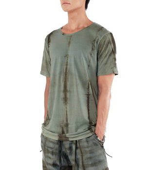 Men looking to left side with his hands in pants pocket, wearing a blue green T shirt dyed with plants. 