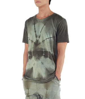 Men looking to the left, with hands in pants pockets, wearing blue green tie dye T shirt 