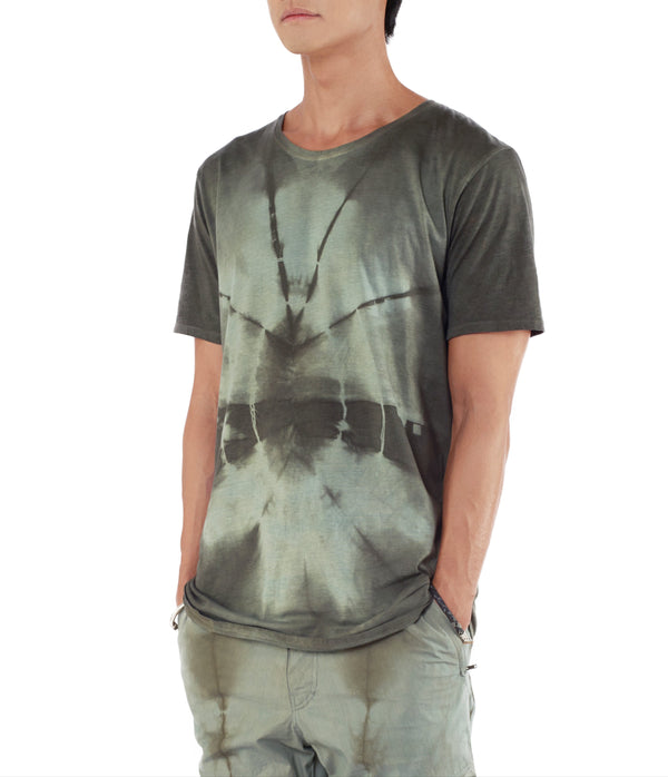 Men looking to the left, with hands in pants pockets, wearing blue green tie dye T shirt 