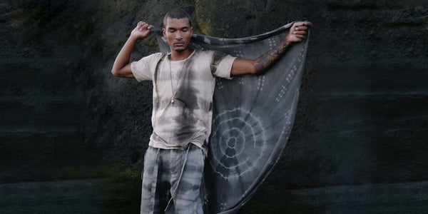 Man holding a large tie dye sarong with a dark background made of rocks 